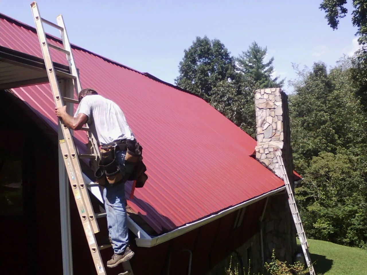 Roofer on a ladder beside a red metal roof and stone chimney — Soffit & Fascia Repair by Roofing Brothers in Danby